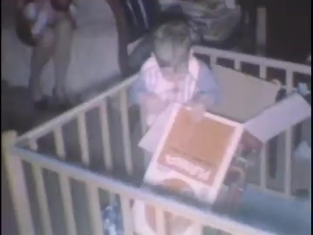 The still captures a young child sitting inside a wooden playpen, intently exploring a cardboard box labeled with a logo. The setting suggests a home environment, with a cozy atmosphere highlighted by soft lighting. In the background, a couple of adults can be faintly seen, indicating a family gathering or casual home moment. The scene embodies a sense of innocence and curiosity, reflecting the joy of childhood.