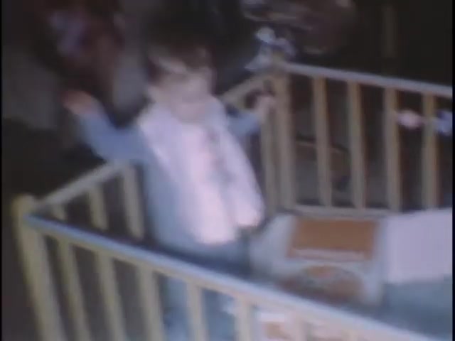 The still shows a young child in a yellow crib, standing with arms raised. The child wears a light blue outfit and a bib. The background is slightly blurred, suggesting a home setting. It captures a moment of innocence and playfulness, likely intended as a cherished memory for Susan.