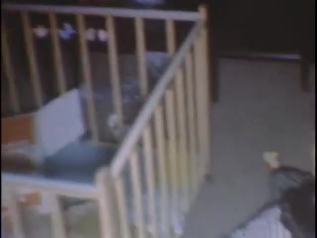 The still features a nursery scene with a wooden crib, possibly showing a child's room. The crib is empty and positioned against a wall, with dim lighting that adds a nostalgic feel. The floor is visible, hinting at a casual home environment. Items around the crib suggest a personal space, likely for a young child, capturing a moment of familial life.