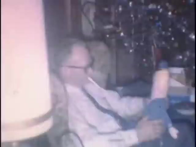 The still depicts an older man seated comfortably in a cozy living room, likely during a festive occasion, given the decorated Christmas tree faintly visible in the background. He is wearing a collared shirt and tie, and is holding a toy, possibly a stuffed animal or doll. There is a gentle glow from a nearby lamp, adding to the nostalgic atmosphere of the home movie.