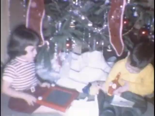 The still features two young children seated on the floor by a decorated Christmas tree. One child, wearing a striped shirt, is playing with an Etch A Sketch, while the other, in a yellow shirt, appears to be engaged with a toy or book. The scene captures a moment of joy and excitement typical of home movie memories during the holiday season.