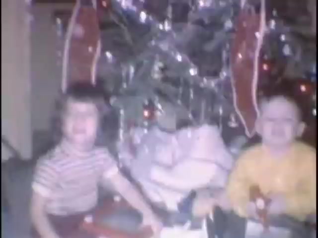 The still features two smiling children seated in front of a decorated Christmas tree. One child wears a striped shirt, and the other is in a yellow sweater. They are surrounded by wrapped gifts and festive decorations, creating a joyful holiday atmosphere.
