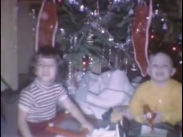 The still features two young children sitting in front of a decorated Christmas tree, surrounded by presents. One child wears a striped shirt, while the other is dressed in a yellow top. The scene captures a festive atmosphere, with ornaments and tinsel adorning the tree, evoking a sense of holiday joy and childhood wonder.