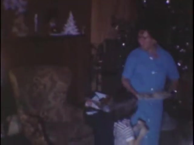 The still depicts a cozy interior scene in the Sisney house, likely during a holiday gathering. A man wearing a blue shirt stands, holding something in his hands, while a child in striped attire playfully interacts around his legs. In the background, a decorated Christmas tree and festive decorations add to the warm, familial atmosphere. The muted color palette and soft focus suggest a nostalgic feel typical of home movies.