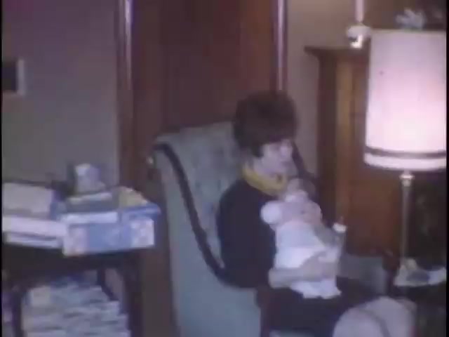The still captures a moment in a cozy living room, featuring a woman seated in a patterned armchair. She is gently holding a baby doll and appears to be engaged in a tender moment, possibly feeding the doll. The room contains a few stacks of magazines or papers on a side table and a lamp that softly illuminates the scene, evoking a warm, nostalgic atmosphere typical of home movies.