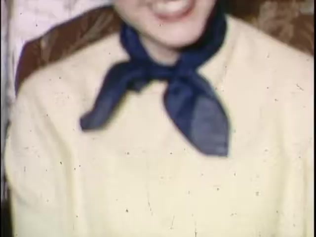 The still features a smiling individual wearing a light-colored sweater and a blue scarf tied around their neck. The image has a vintage aesthetic, typical of 16mm film, with noticeable grain and a slightly blurred focus. The background appears to be a patterned surface, adding to the nostalgic feel of the image.