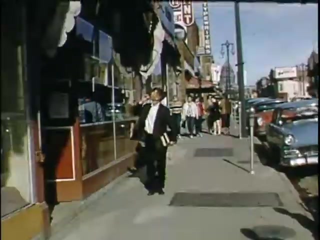 The still depicts a bustling street scene from the mid-20th century. A man in a suit, holding books, walks along the sidewalk, while a crowd can be seen in the background, alongside vintage cars parked on the street. The storefronts are adorned with awnings, and signage is visible, suggesting a vibrant commercial area. The bright colors typical of Kodachrome film enhance the nostalgic ambiance.