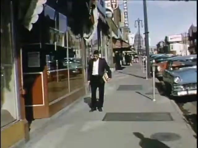 The still shows a man walking along a sidewalk, dressed in a black suit with a white shirt, holding a book. The scene captures a bustling urban setting with shops visible, including a neon sign in the background. Classic cars are parked along the street, suggesting a mid-20th century timeframe. The overall atmosphere appears lively and dynamic.