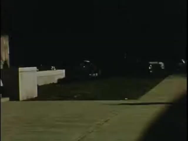 The still captures a dimly lit street scene, highlighting a wide sidewalk with a concrete edge. Cars are parked along the street, partially visible in the shadows. The overall atmosphere is quiet and somewhat mysterious, likely set during evening or night. The film’s vintage quality, characteristic of Kodachrome, imparts a nostalgic feel to the image.