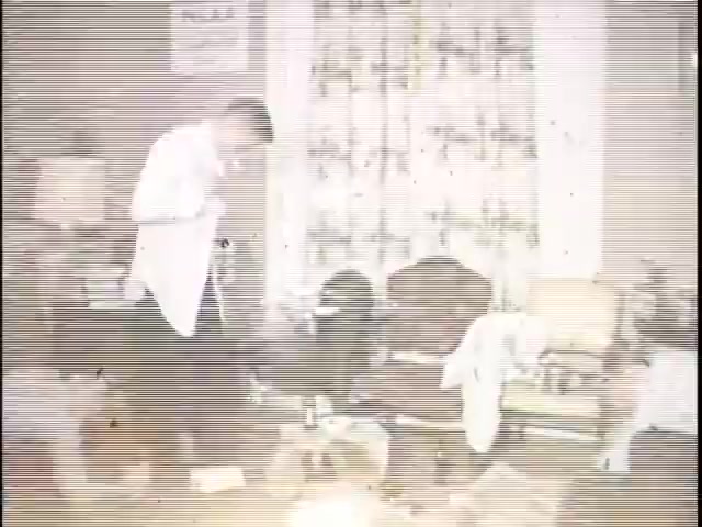 The still depicts a man in a white shirt standing in a cluttered room, appearing to examine or work with something in his hands. The background features patterned curtains and various items scattered across the furniture, suggesting a domestic setting with an unkempt appearance. The image has a vintage look, characteristic of 16mm film, with visible grain and lines.