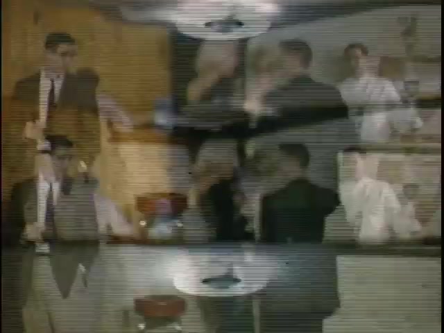 The still captures a vibrant scene typical of vintage film, featuring a group of individuals in a social setting, possibly a gathering or a party. The image exhibits layered visuals, suggesting overlapping moments or dialogue, with characters dressed in formal attire. The color tones reflect the nostalgic quality of Kodachrome film, lending a warm atmosphere to the interaction. The background hints at a domestic space, adding to the intimate and casual vibe of the gathering.