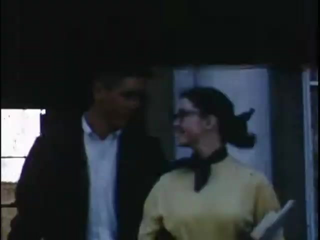 The still features a young man and woman walking closely together, engaged in a lighthearted conversation. The woman wears a yellow top accented with a black scarf, and the man is dressed casually. Their expressions suggest familiarity and enjoyment, set against a backdrop that appears to be a building or structure, with soft, muted colors characteristic of vintage Kodachrome film.
