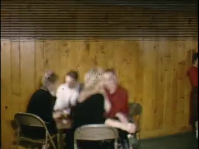 The still depicts a group of individuals sitting at folding chairs in a casual setting, likely a gathering or event. They appear engaged in conversation, with some figures slightly obscured. The background features wooden paneling, creating a warm, rustic atmosphere. The image exhibits a vintage aesthetic typical of 16mm film.