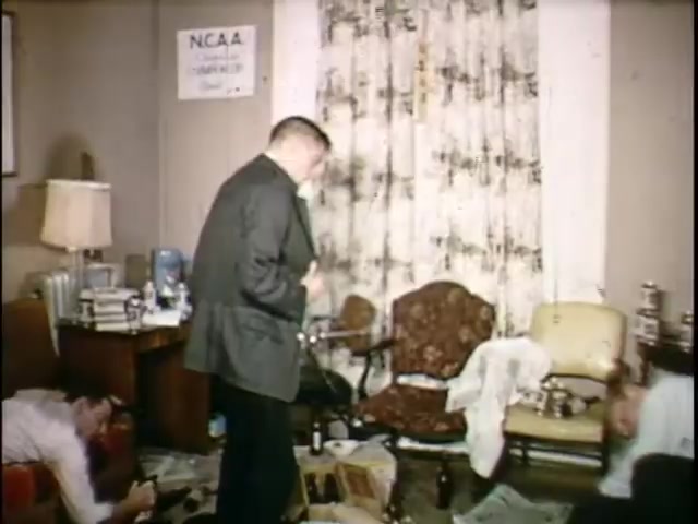 The still features a cluttered room with two men. One man, dressed in a suit, stands and appears to be adjusting his clothing. The other man, in a white shirt, is sprawled on the floor. Surrounding them are scattered newspapers, empty bottles, and unkempt furniture, highlighting a disheveled environment. A curtain with a floral pattern and an NCAA sign on the wall adds context to the setting.