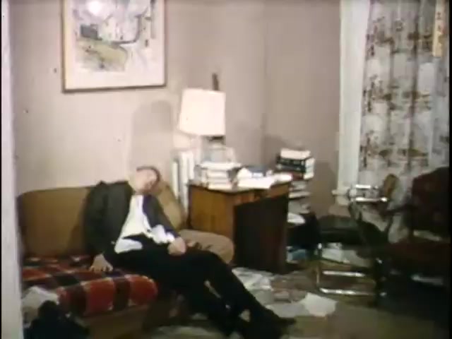 The still depicts a disheveled living room featuring a man slumped on a couch, suggesting a state of exhaustion or despair. The surroundings are cluttered, with stacks of books and scattered debris, creating a chaotic atmosphere. A lamp and artwork are visible, contributing to the domestic setting. The overall mood conveys a sense of disarray or distress.