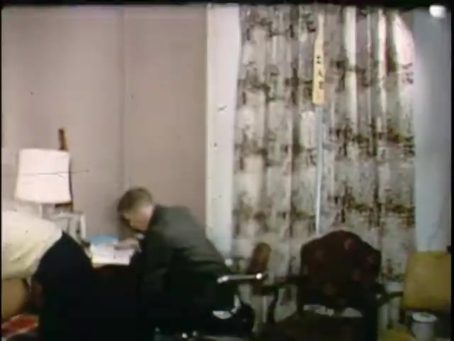 The still from 'kodachrome 20140420' features two men in a room with a modest decor. One man, dressed in a suit, is seated and focusing on papers on a table, while the other man, in a lighter shirt, is leaning down, possibly organizing or retrieving something. The room has patterned curtains and simple furnishings, evoking a mid-20th century aesthetic. The lighting is soft, contributing to a casual atmosphere.