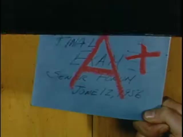 The still features a hand holding a blue piece of paper displaying a large red 'A+' grade. It is labeled 'FINAL EXAM - SENIOR FORUM' with the date 'JUNE 12, 1986' written in smaller text. The background appears to be a wooden surface, suggesting a casual or informal setting.