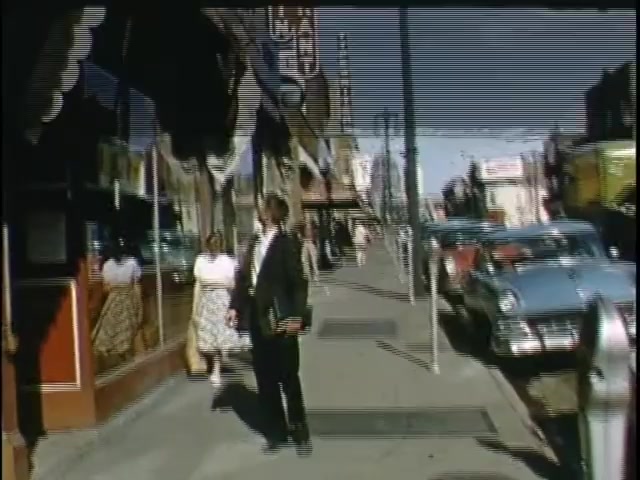 The 16mm still from 'kodachrome 20140420' captures a bustling street scene. A man in formal attire stands on the sidewalk, looking toward a storefront where two women in dresses are reflected in the window. The street is lined with vintage cars, showcasing a mid-20th-century setting. The overall ambiance is nostalgic, hinting at a lively community atmosphere from the past.