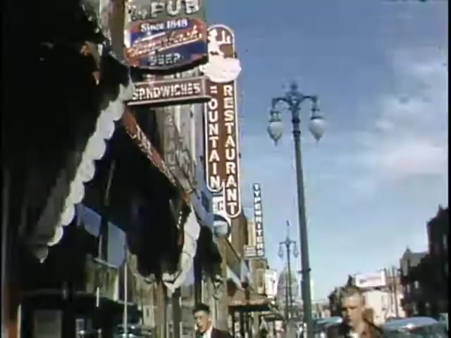 The still features a lively street scene with various colorful signs for restaurants and pubs, showcasing a vintage atmosphere. The architecture reflects a mid-20th century aesthetic, with a sunny sky overhead. Pedestrians can be seen walking along the sidewalk, contributing to the bustling vibe of the area. The overall image captures a nostalgic urban environment.