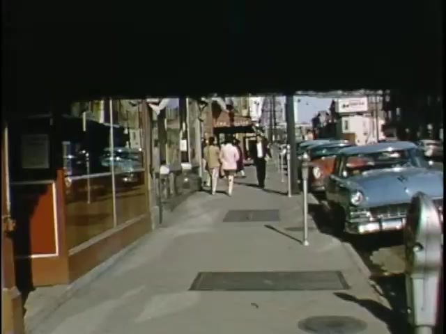 The still depicts a vibrant street scene featuring pedestrians walking along a sidewalk. In the background, storefronts and parked vintage cars, including several blue models, are visible. The colors are vivid, characteristic of Kodachrome film, suggesting a lively atmosphere. The image conveys a sense of everyday life, likely from the mid-20th century, with people casually interacting in an urban setting.