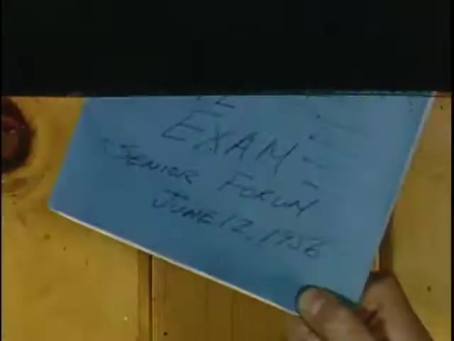 The still features a hand holding a blue card that reads 'EXAM: Senior Forum June 12, 1976.' The background shows a wooden surface, likely a wall, adding a rustic feel to the image. The focus is on the card, emphasizing the significance of the exam context.