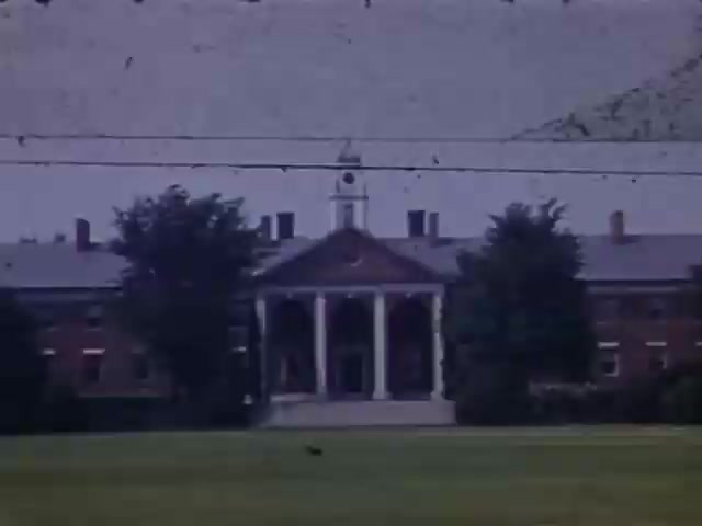 The still features a historic building characterized by classical architecture, prominently displaying a portico with columns. Surrounding the structure are large trees and a well-manicured lawn, contributing to a tranquil atmosphere. The overall color palette suggests warmth typical of Kodachrome film, with slight artifacts indicating the age of the image, likely captured between 1941 and 1943. Background details include distant hills, adding depth to the scene.