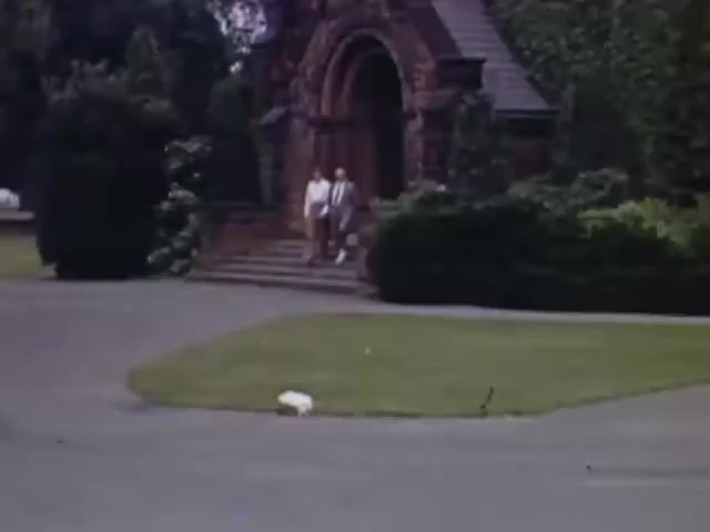 The still from 'Jack #2' features two individuals walking down the steps of a stone building surrounded by lush greenery. The scene captures a serene moment, likely in a residential area, with a well-maintained lawn in the foreground. The Kodachrome film emphasizes vibrant colors, typical of the early 1940s. The overall atmosphere is nostalgic, reflecting a leisurely day outdoors during that era.