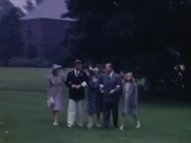 The still depicts a group of six individuals walking together in a lush, green setting, likely a garden or park. They are dressed in formal attire typical of the early 1940s, with two women in elegant dresses and hats, while the men wear suits. A sense of camaraderie and celebration is palpable, hinting at a special occasion. The color tones are characteristic of Kodachrome film, adding a nostalgic quality to the scene.