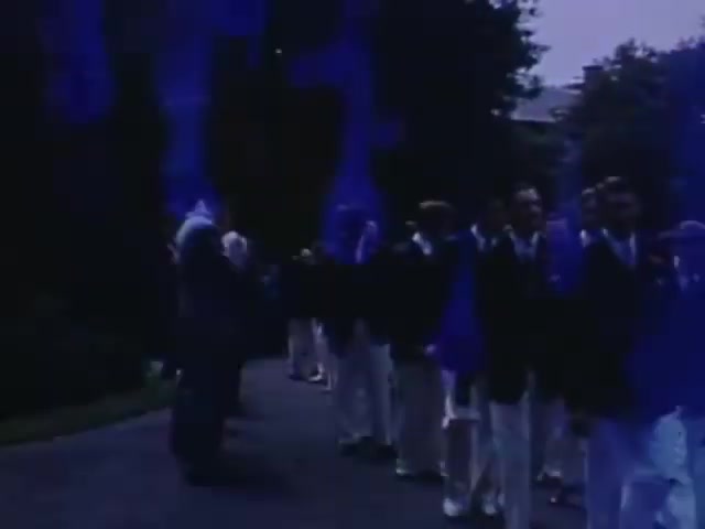 The still from 'Jack #2' features a gathering or procession, capturing several well-dressed individuals in formal attire. The scene seems to take place in a lush outdoor setting, with foliage in the background. The color palette shows a distinct bluish tint, which might be a characteristic of Kodachrome film from that era. The overall mood appears formal and celebratory, typical of events from the early 1940s.
