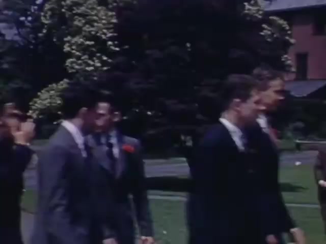 The still from 'Jack #2' features a group of young men dressed in suits, walking together in a park-like setting. Some are wearing boutonnieres, and they appear engaged in conversation. The background includes lush greenery and a building, suggesting a social or celebratory occasion typical of the early 1940s. The Kodachrome film captures vibrant colors characteristic of that era.