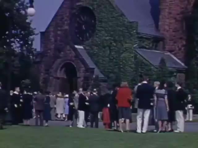 The still captures a gathering outside a stone church, characterized by its ivy-covered facade and prominent clock. Attendees, dressed in formal attire typical of the early 1940s, are mingling on the lawn. The scene conveys a sense of occasion, possibly a wedding or memorial, reflecting the social customs of the time. The vibrant colors suggest the use of Kodachrome film, enhancing the cheerful mood of the gathering.