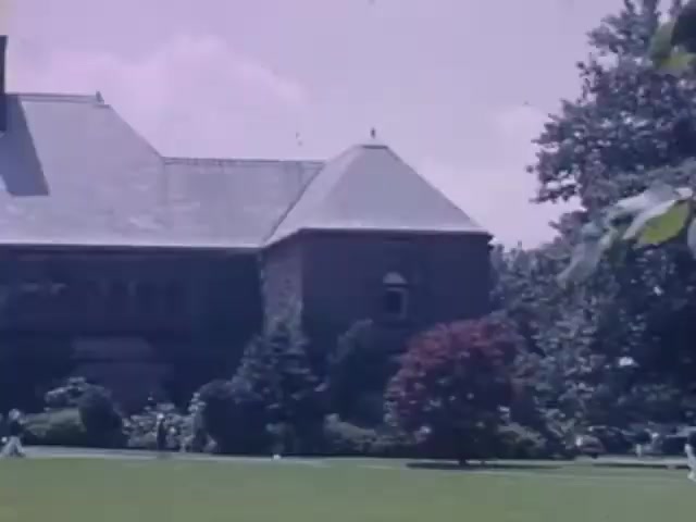 The still from 'Jack #2' features a large, stately building with a distinctive roof, set in a lush green landscape. Surrounding the structure are well-maintained gardens with flowering shrubs and trees. The scene captures a sunny day, contributing to a vibrant color palette typical of Kodachrome film from the early 1940s. There are a few figures in the background, suggesting leisurely activity in the outdoor setting.