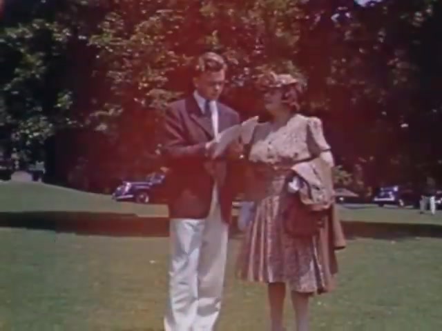 The still from 'Jack #2' features a young man in a blazer and light pants standing outdoors, engaged in conversation with a woman dressed in a floral-patterned dress and a hat. They appear to be reading from a piece of paper while surrounded by a verdant park setting. Classic cars are parked in the background, suggesting a mid-20th century ambiance typical of Kodachrome film from the early 1940s. The colors display the vividness characteristic of the film medium during that era.