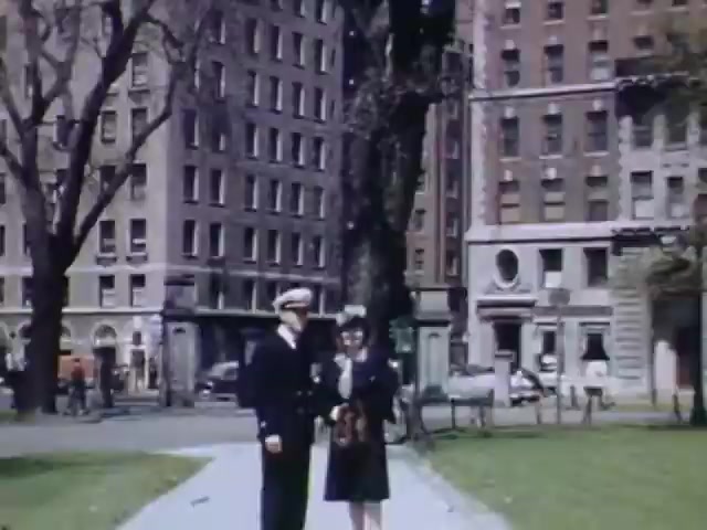 The still from 'Jack #2' showcases a young naval officer in uniform standing next to a woman dressed in 1940s fashion. They are positioned on a pathway in a park with large trees, surrounded by mid-century urban buildings. The image is vibrant, capturing the essence of city life during the early 1940s, likely around the time of World War II. The colors, characteristic of Kodachrome film, enhance the nostalgic atmosphere.