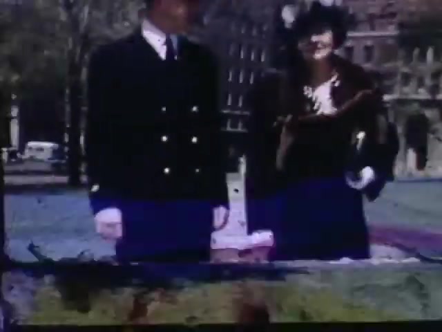 The still from 'Jack #2' features two individuals, a man in a military uniform and a woman in formal attire, standing together outdoors. The scene appears vibrant due to the use of Kodachrome film, which captures rich colors typical of the early 1940s. The background hints at an urban setting with old architecture, suggesting a moment captured during a time of social gathering. The image exhibits signs of age, with slight discoloration and wear characteristic of vintage film.