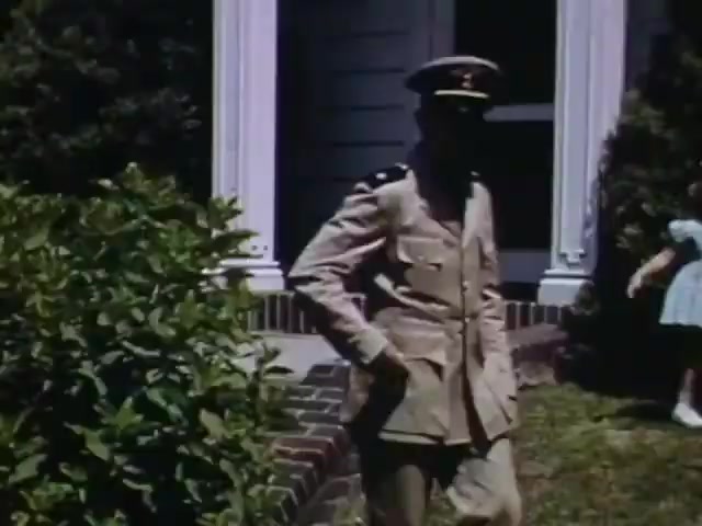 The still features a military figure in a light-colored uniform and cap, standing at the edge of a garden. He appears to be in a relaxed stance, with one hand in his pocket. In the background, there's a house with a white porch, and a child wearing a blue dress is partially visible. The Kodachrome color palette highlights the vivid hues typical of early 1940s film, suggesting a warm, nostalgic atmosphere. This scene reflects the era's familial and patriotic themes.