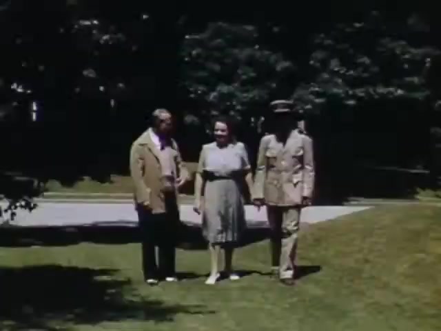The still shows three individuals—two men and a woman—walking together on a lawn, likely in a park or a suburban setting. One man is dressed in casual attire, while the other wears a military uniform, suggesting a wartime context. The woman, dressed in a vintage dress, appears engaged in conversation with both men. The scene is bathed in bright, natural light, characteristic of Kodachrome film from the early 1940s.