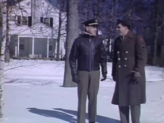 The still from 'Jack #2' captures two men outdoors in a snowy setting during the early 1940s. One man is dressed in a military uniform with a leather jacket and cap, suggesting a role in the armed forces, while the other wears a long, double-breasted overcoat. They appear to be engaged in a conversation, with a residential house visible in the background, typical of that era. The color palette reflects the vibrant tones of Kodachrome film, enhancing the nostalgic feel of the scene.