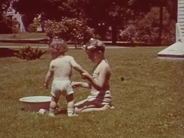 The still from 'Connie & Patty #2' features two children playing outdoors on a grassy lawn. One child is sitting and interacting with a younger child, who is standing and appears to be exploring a small basin of water. The older child is likely wearing a swimsuit, suggesting a warm, sunny day. The setting feels relaxed and playful, indicative of a carefree childhood, possibly during the mid-20th century, around the WWII timeframe as suggested. The colors and film stock point to a domestic scene typical of home movies from that era.