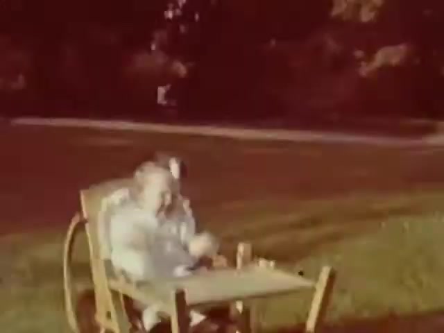 The still shows two children sitting in a high chair, likely outdoors on a sunny day. The film's color suggests a warm, nostalgic tone, typical of mid-20th century home movies. Given the style and medium (Ansco film), your guess of the WW2 timeframe seems plausible. The children's clothing and the setting evoke an era where family and outdoor leisure were commonly captured in film.