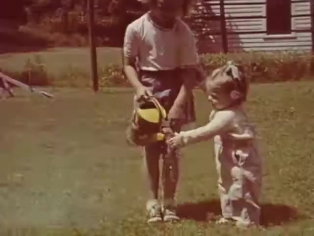 The still depicts two young girls outdoors, one likely older and holding a toy lawnmower while the younger girl, dressed in overalls, attempts to use it. The setting appears to be a backyard with a grassy area and a house in the background, suggesting a domestic scene typical of the mid-20th century. The use of Ansco film implies it may have been shot during or shortly after WWII, during a time when color film was becoming more common in home movies. The clothing style and the playful activity further support this timeframe.