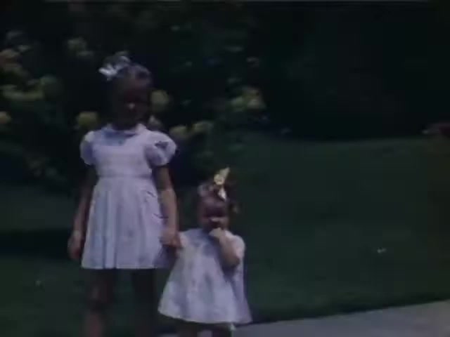 The still from 'Connie & Patty #2' features two young girls, one older and one younger, both dressed in light-colored dresses. The older girl is holding hands with the younger one, who has a bow in her hair. The background suggests a suburban setting with greenery, typical of the mid-20th century. The Ansco film suggests it may be from the late 1940s, possibly around the World War II timeframe, though exact dating is difficult without more context.