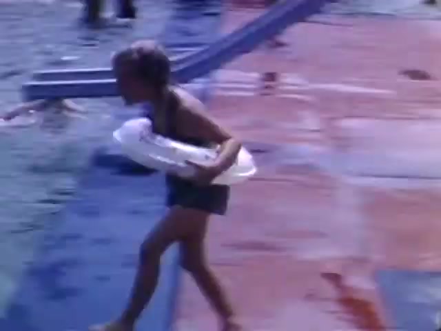 The still depicts a child running by a swimming pool, holding a flotation device. The brightly colored ground indicates a cheerful summertime setting. Given the Ansco film type and the playful nature of the scene, it likely dates to the mid-20th century, possibly around the World War II era. The simplicity of the child's attire and the family-friendly activity suggest a focus on leisure during a time when such moments were cherished amidst larger social tensions.