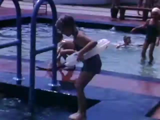 The still from 'Connie & Patty #2' depicts a group of children enjoying a day at the pool, surrounded by classic 1940s swimwear. One child is climbing out of the pool with a floatation device, while others swim in the background. The Ansco film stock suggests a date around the WWII timeframe, as the aesthetic and style of the scene align with leisure activities typical of that era. Overall, it captures a nostalgic, carefree moment in childhood.