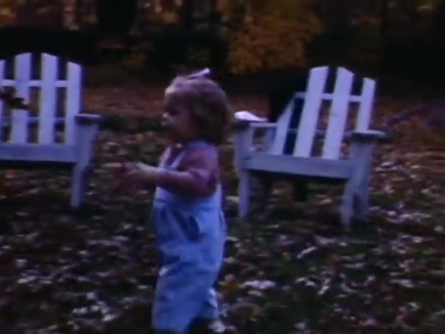 The still depicts a young child in a playful moment, wearing blue overalls and a light top, suggesting a carefree outdoor setting. The background features two white wooden chairs and a landscape with fallen leaves, indicative of autumn. Given the Ansco film stock and the style of children's clothing, the film likely originates from the late 1930s to early 1940s, possibly around the World War II timeframe. The warm color palette and homey atmosphere evoke a nostalgic, family-oriented scene from that era.