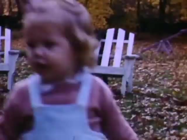 The still depicts a young child with blonde hair in a blue romper, playing outdoors amidst autumn leaves. In the background, there are white wooden chairs and hints of fall foliage, suggesting a natural setting. The film likely captures a nostalgic moment from childhood, possibly around the WWII era, given your context about the time frame and the use of Ansco film. The warm colors and soft focus evoke a sense of simplicity and innocence typical of home movies from that period.