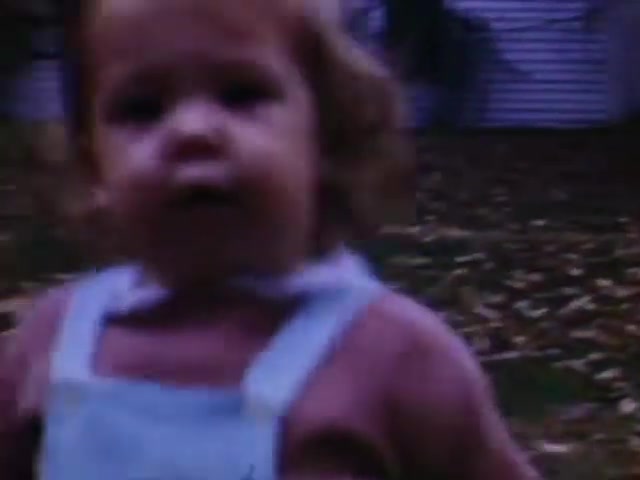 The still appears to show a young child, possibly a toddler, with curly hair, wearing a light blue outfit and a pink top, amidst what looks like a backyard setting with fallen leaves. The coloring and quality suggest it was shot on 16mm Ansco film, which was more common in the early to mid-20th century. Given the style of clothing and the film type, your guess of a WW2 timeframe seems plausible, likely dating it to the 1940s.