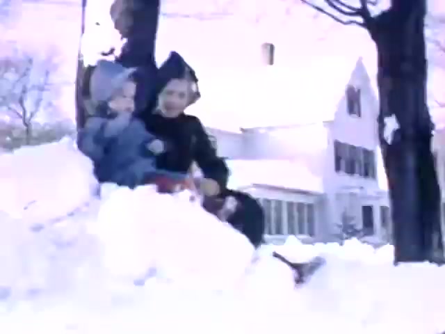 The still from 'Connie & Patty #2' features two children playing in the snow, suggesting a winter setting. They are dressed in warm clothing typical of the era, which may suggest a time frame around World War II. The background shows a house with a classic architectural style, likely indicative of mid-20th century American homes. The film’s use of Ansco film stock, rather than Kodak, also supports this timeframe, suggesting it’s from the late 1930s to early 1940s.