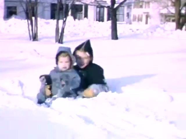 The still from 'Connie & Patty #2' depicts two children sitting in a snowy landscape, suggesting a winter setting. They are bundled up in warm clothing, typical of cold weather. The film stock is Ansco, which was popular in the mid-20th century. Based on the clothing and the context, it likely dates around the World War II era, possibly the early to mid-1940s. The serene winter scene captures a playful moment during a time of significant historical events.