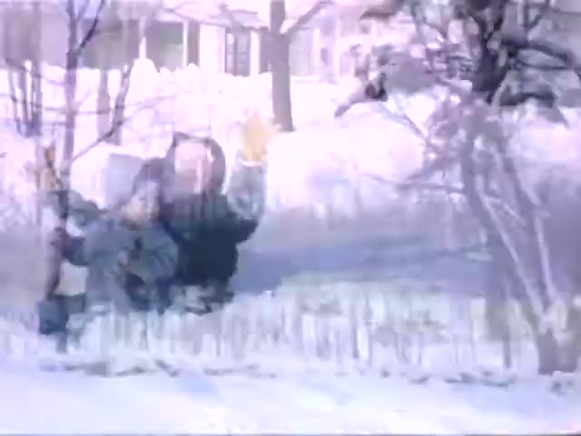 The still from 'Connie & Patty #2' captures a snowy outdoor scene featuring two individuals. One person appears to be lying in the snow with arms outstretched, possibly in play or in a casual pose. The background suggests a residential area with trees and a house partially obscured by snow. The Ansco film quality hints at a time when color film was less common, potentially placing the scene in the mid-20th century, possibly around the WWII era.